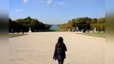 Woman with a black backpack walking through a sunny formal garden towards a distant lake and trees.