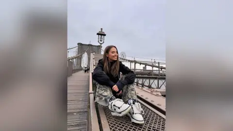 EvelynMills sits on a metal walkway on a bridge, wearing a black long-sleeve shirt, camouflage pants, and white sneakers.