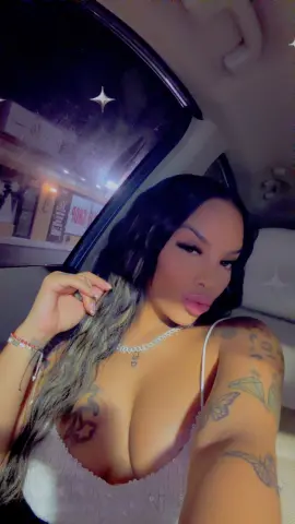 Tattooed woman in a white sequin top posing inside a car at night with a glamorous mood.