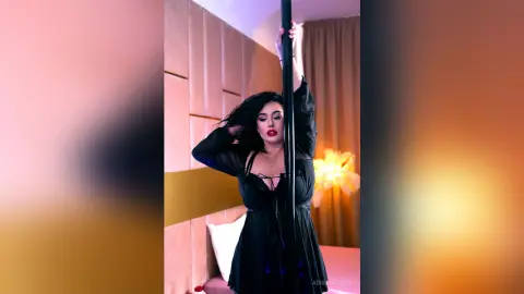 AdiraMaeven poses with a black dance pole, wearing a sheer black long-sleeve top and dark hair styled down.