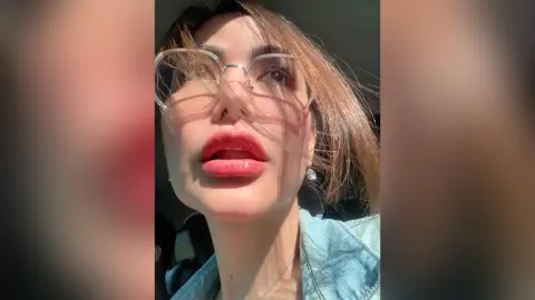 Close-up portrait of a woman with glasses and red lipstick looking up in soft natural sunlight.