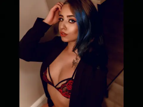 Young woman with blue hair wearing a black blazer and red lace bra in a warm indoor setting.