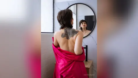 IveteDurand poses with her back to the camera, showing her dark hair and large back tattoo while wearing a bright pink silk robe in a bathroom setting.