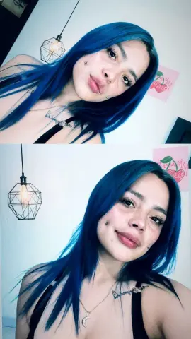 Smiling young woman with long blue hair wearing a black tank top and silver necklace in a bright room.