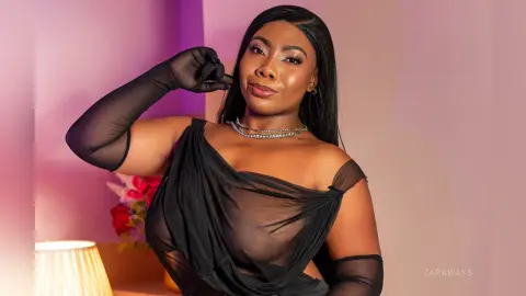 Elegant woman in a sheer black evening dress and long gloves posing against a soft pink studio background.