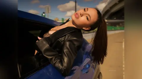 DelanaGrace leans out of a blue car window wearing a black leather jacket and black lace bra, her hair in a long ponytail.