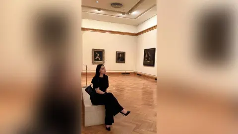 Elegant woman in a black outfit sitting in a quiet art museum gallery surrounded by classical paintings.