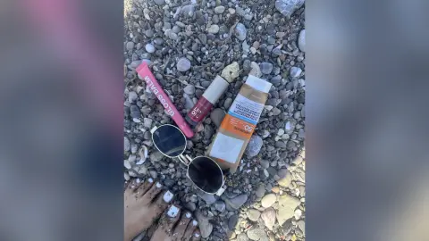 Pink lip gloss, sunblock, and sunglasses resting on beach pebbles under bright summer sunlight.