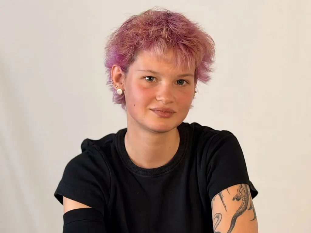 Young woman with short pink hair and black t-shirt smiling against a plain white background.