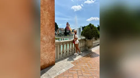 DemmiLovat — DemmiLovat stands on a stone balcony wearing a white top and shorts, looking out at a large fountain under a blue sky.