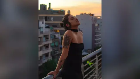 Tattooed woman in a black dress enjoying a peaceful sunset on an urban city rooftop balcony.