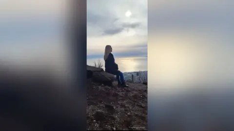 Blonde woman in a dark jacket sitting on a rocky cliff overlooking a vast ocean under a cloudy sky.