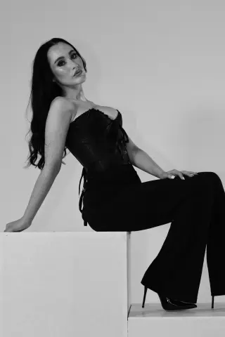 Elegant woman in a black corset and trousers sitting on a white pedestal in a minimalist studio setting.