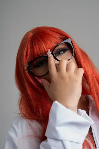 Woman with bright orange hair and black glasses wearing a white shirt in a minimalist studio setting.