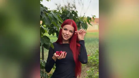AriaVex smiles while holding red strawberries in her hands, wearing a long-sleeved black top against a leafy green background.