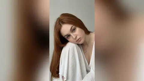 Redheaded woman in a white textured blouse posing with a serene expression against a neutral background.