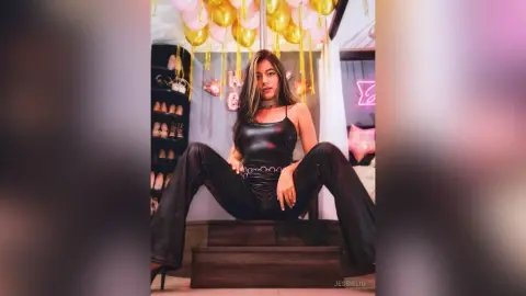 JessieLiu wears a black leather bodysuit and sits on a dark wooden stool, posing with her legs spread wide.