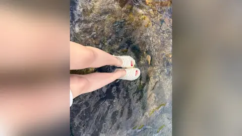 EmilyGiffin dangles her bare legs over clear, shallow running water with her white sandals resting near the rocks.