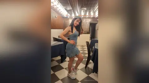 Smiling young woman in a denim romper posing in a modern restaurant with checkered floors.