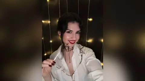 Smiling young woman in a white button-down shirt posing against a dark background with fairy lights.