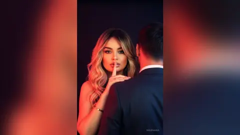 Glamorous blonde woman in elegant evening wear posing intimately with a man in a black suit against a red backdrop.