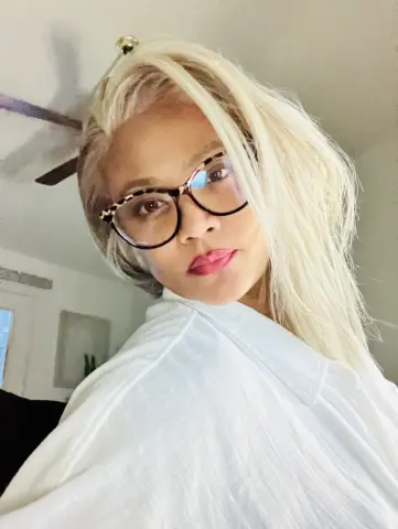 Blonde woman in glasses and a white linen shirt smiling in a bright indoor setting.