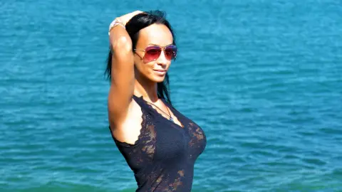 Smiling woman in a black lace tank top and sunglasses relaxing by the bright blue ocean water.