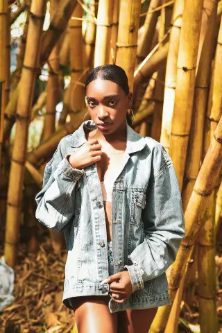 Young Black woman in denim jacket posing against a golden bamboo forest background in warm sunlight.