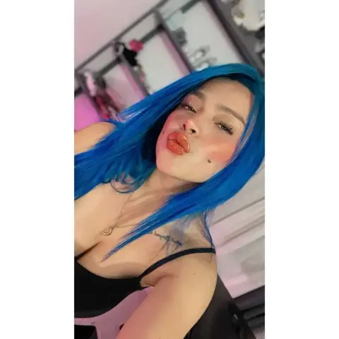 Young woman with bright blue hair in a black top posing in a bedroom setting.