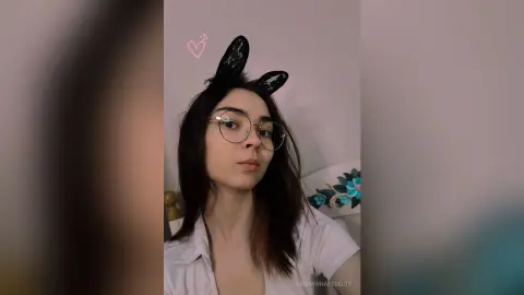 Young woman with glasses and black bunny ears wearing a white shirt in a cozy indoor setting.