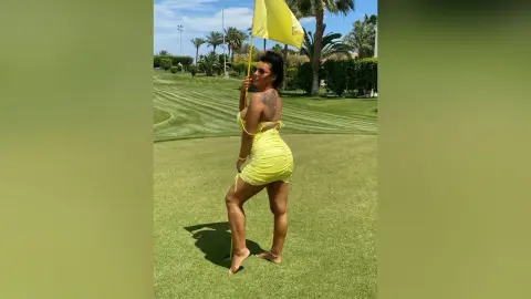 MissyJolie stands on a green golf course wearing a tight yellow mini dress and sunglasses.