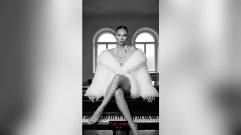 Elegant woman in a white faux fur coat and pearl necklace sitting on a piano in a glamorous black and white setting.