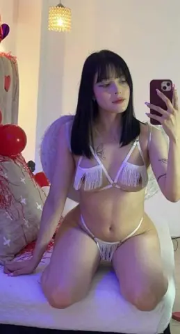 Young woman in white fringe bikini sitting on a bed in a cozy, dimly lit bedroom setting.