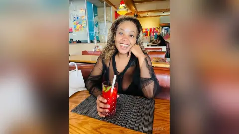 Smiling woman in a black sheer blouse enjoying a red berry drink at a cozy cafe table.