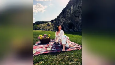 Woman in a white floral dress sitting on a picnic blanket near a rocky mountain under a sunny sky.