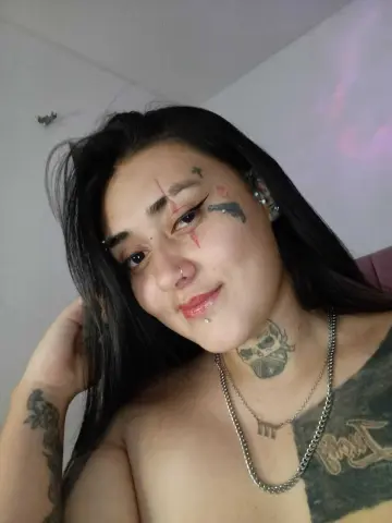 Smiling woman with facial tattoos and silver necklaces posing in a casual indoor setting.