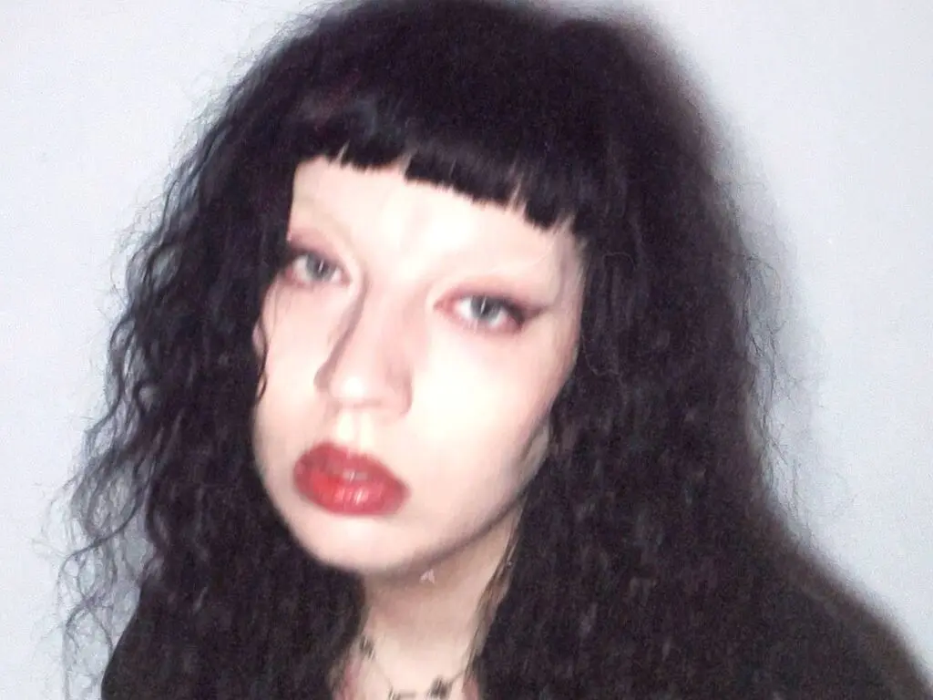 AnnaDark has long, wavy black hair with blunt bangs and red lipstick, looking directly at the camera against a plain white background.