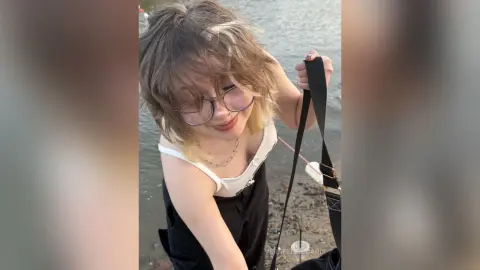 VernellZiccardi with messy blonde hair and glasses, wearing a white tank top and black straps, posing outdoors by the water.