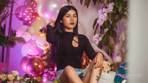 Young woman in a black long-sleeve top sitting amidst pink balloons and flowers in a dreamy, soft-lit setting.