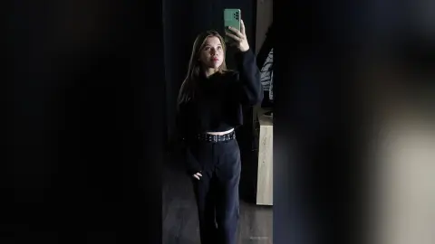 Young woman in a black cropped sweatshirt and trousers taking a moody mirror selfie in a dimly lit room.