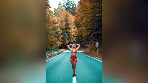 AmelieSkylar stands on a paved road surrounded by autumn trees, wearing red lace lingerie and holding her hands behind her head.