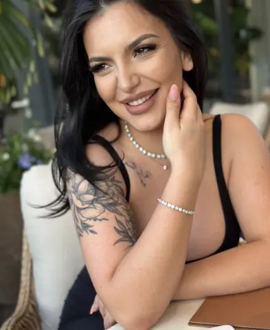 NoraSin smiles while resting her hand against her face, showing off her long black hair, black tank top, and silver jewelry.