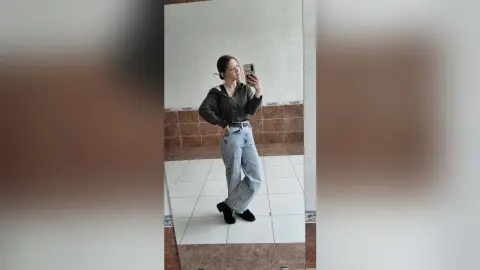 Young woman in a dark hoodie and light wash jeans taking a mirror selfie in a modern tiled room.