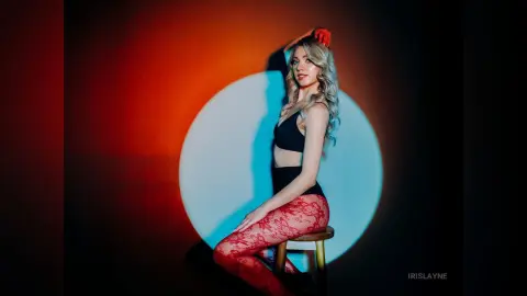 IrisLayne poses sideways in a black bra and high-waisted bottoms with red lace patterned tights against a bright circular light.