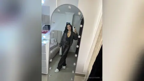 Stylish woman in a black leather jacket and trousers taking a selfie in a lighted vanity mirror.