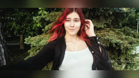 Smiling young woman with red hair wearing a white tank top and black jacket in a lush green garden setting.