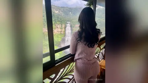Woman in a pink ribbed lounge set looking out a window at a scenic jungle waterfall view.