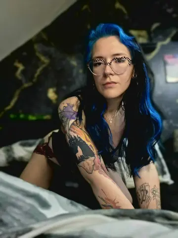 Harley_MoonX with blue hair and glasses poses in a black top, showing off her colorful arm tattoos while leaning on a bed.