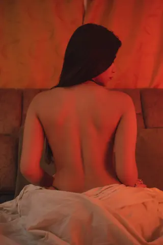 Back view of a woman in white fabric sitting in a moody, red-lit room.