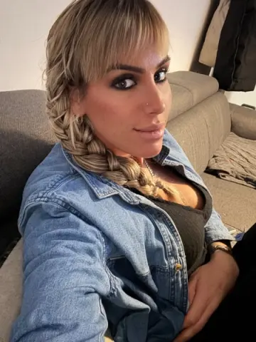 Blonde woman with braided hair wearing a denim shirt sitting on a grey sofa in a cozy indoor setting.